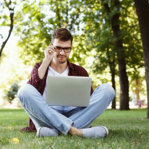 Man working outdoors on laptop. Preparing for exams, sitting on grass at university campus park. Technology, communication, education and remote working concept, copy space
