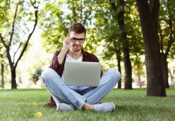 Man working outdoors on laptop. Preparing for exams, sitting on grass at university campus park. Technology, communication, education and remote working concept, copy space