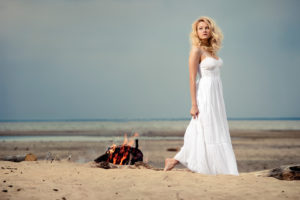 A barefoot woman wearing a white dress on the beach near an open fire.